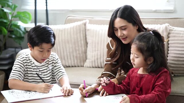 Asian mother watching her cute son and little daughter drawing pictures together in living room at home.