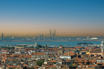 Obraz premium Historic Venice rooftops and a calm lagoon contrast with distant industrial smokestacks under a hazy sky. A powerful visual for environmental campaigns on urban pollution and sustainable futures.