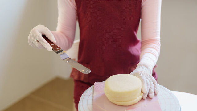 Asian baker wearing gloves spreading frosting on pink cake layer with palette knife on turntable