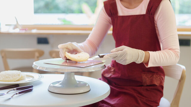 Asian woman in burgundy apron spreading frosting on white cake layer using rotating stand in bright kitchen