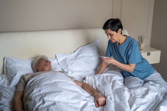 Concerned senior woman checking temperature level with thermometer of sick partner lying in bed, worried elderly female taking care of weak husband suffering from flu, health issues at older age