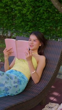 Asian woman in swimsuit sitting on woven bamboo chair reading book