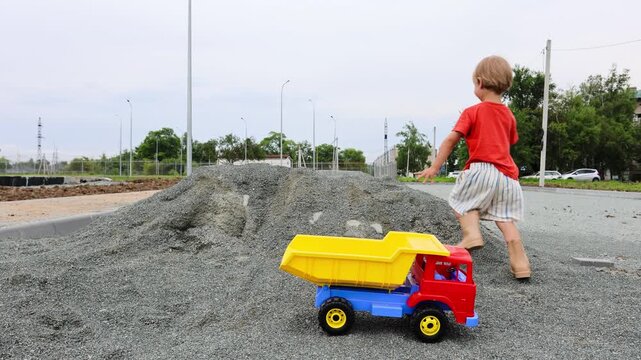 A 4-year-old boy wearing shorts and rubber boots plays with a large toy dump truck on a summer day. He loads the truck bed with gravel using his hands. Active outdoor play of a child