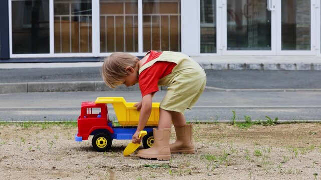 A blond 4-year-old boy wearing a khaki short romper and beige rubber boots plays in the backyard on a sunny summer day. The child uses a yellow plastic shovel to load sand into a toy dump truck 