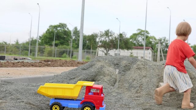A 4-year-old boy wearing shorts and rubber boots plays with a large toy dump truck on a summer day. He loads the truck bed with gravel using his hands. Active outdoor play of a child