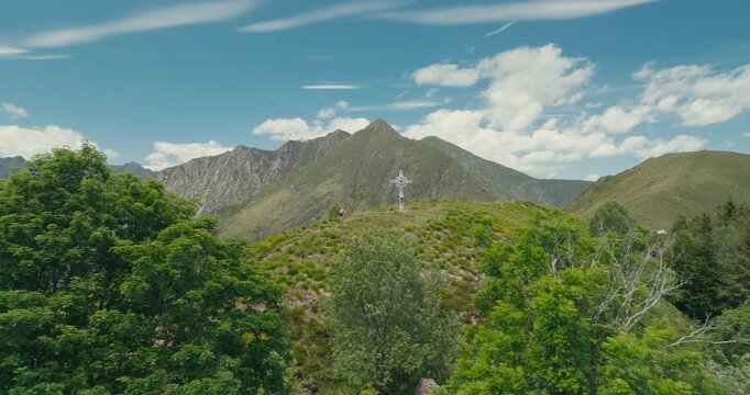 Drone flies over trees toward a summit cross as a female hiker walks the final meters to the peak, surrounded by breathtaking mountain views beneath a clear blue summer sky.