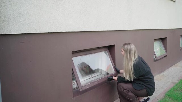 Long-haired homeowner in dark clothing tests an old tilted basement window frame, checking for damage and planning repair or replacement.