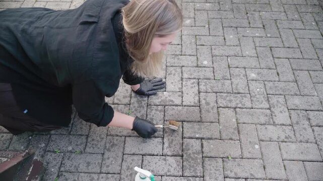 Black-gloved DIY cleaner scrubs and sprays a stained brick patio, battling weeds and grime between the paving joints.