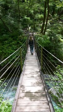 A female traveler with a backpack walks across a wooden bridge over a turquoise river in the lush green Tolmin Gorge. Perfect for travel vlogs and wellness content.