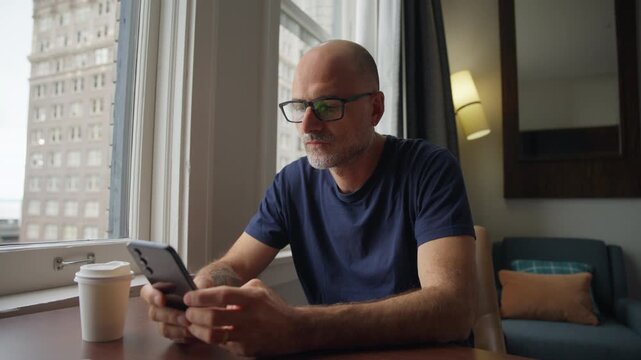 Man puts on eyeglasses, smiles while using smartphone, and checks messages at a home office table with coffee cup and city view.