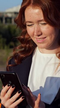 Smiling young woman with beautiful red curly hair and freckles scrolling and typing on her smartphone while enjoying the sunny weather in a city park, showing modern digital communication