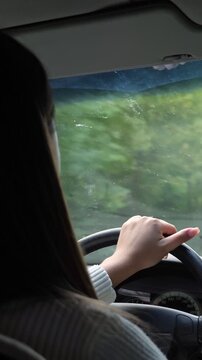 Unrecognizable woman driving her car on a scenic road trip through the countryside, with her hands holding the steering wheel and green trees visible through the windshield