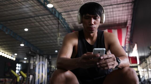 Muscular Asian man listening to music on his smartphone in a gym.