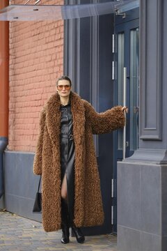 A fashionable woman in a long brown fur coat and sunglasses walks out of a colorful urban office building