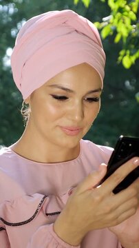 Young muslim woman wearing a pink hijab happily texting on her smartphone while standing in a park, enjoying the sunny day and modern technology for communication and social networking