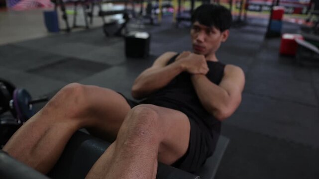 Muscular Asian man doing intense ab crunches at the gym. Athletic young man performing abdominal crunches on a bench. Intense core workout for building strong six-pack abs and improving fitness.
