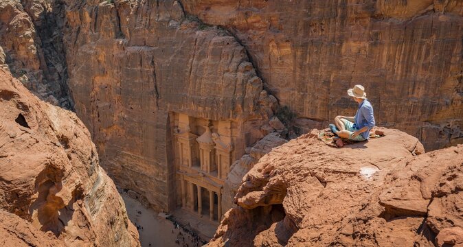 Tourist with sun hat sits on rocks and looks from above into the canyon Siq, Pharaoh's treasure house beaten into rock, facade of the treasure house Al-Khazneh, Khazne Faraun, mausoleum in the Nabataean city Petra, near Wadi Musa, Jordan