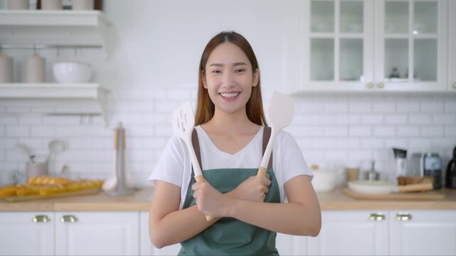 Happy young asian woman wearing apron and standing in kitchen room. Beautiful female smiling and looking at camera feeling confident