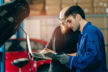 Two male mechanics working together in garage, one holding laptop and other checking clipboard,...