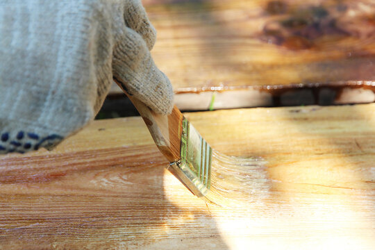 Gloved hand brushing wood preservative onto a wooden plank outdoors. Home renovation, DIY woodworking, timber maintenance and protective coating concept.