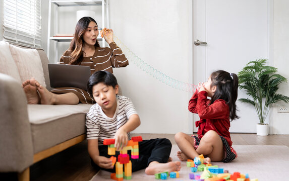 Cheerful Asian mother playing slinky toy with kids while working on laptop at home