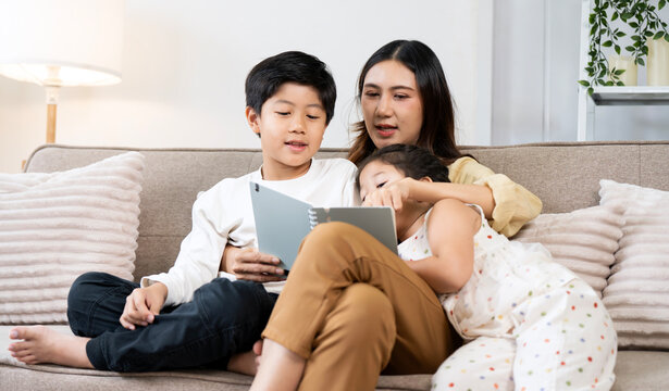 Asian mother reading a book with her son and daughter on sofa at home