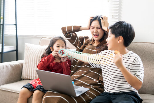 Stressed Asian mother holding head in frustration while kids playing rowdy with toy over her on sofa