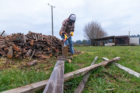 Low angle view of a hispanic man splitting a log with an axe next to a large firewood pile