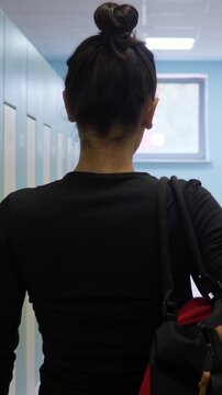 Rear view of a fit woman with a bun hairstyle wearing sportswear and carrying a gym bag, walking down a hallway with lockers before her workout at the fitness club or school