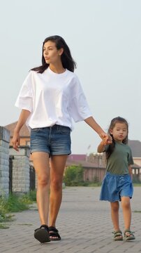 Happy family time as a young mother and her cute asian daughter enjoy walking together holding hands on a suburban street during a sunny day, showcasing love, care, and a strong family bond