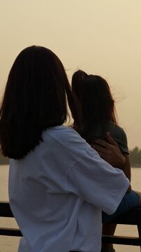 Loving mother gently embracing her daughter while sitting on a railing, both enjoying the tranquil view of the sunset over the water, creating a tender and peaceful family moment outdoors