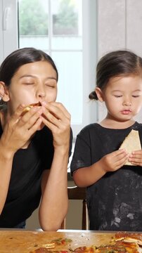 Affectionate mother and her cute little daughter enjoying freshly baked homemade pizza together in the kitchen, sharing a happy family moment while eating and laughing after a fun cooking session