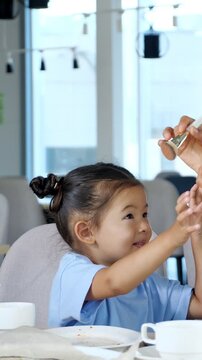 Charming little korean girl with space buns hairstyle smiling happily as a makeup artist applies blush to her cheeks with a brush, enjoying the cosmetic procedure at a table in a bright cafe