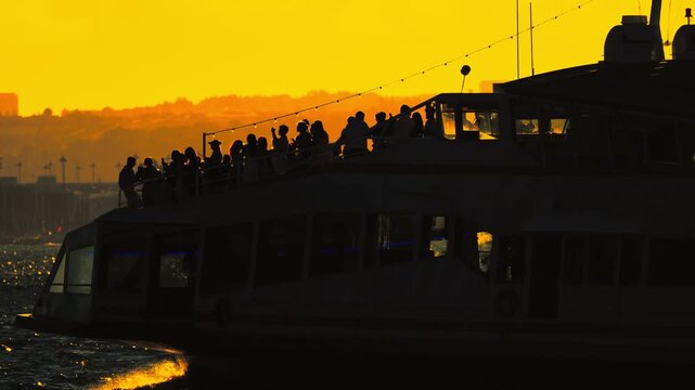 Group of people enjoying sunset views from the upper deck of a ferry boat, silhouetted against a vibrant orange sky over the water