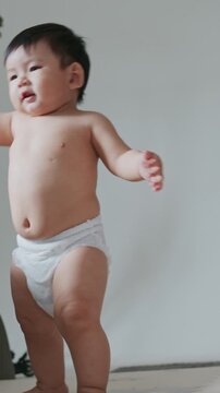Happy Asian baby boy in diaper learning to walk with arms raised for balance.