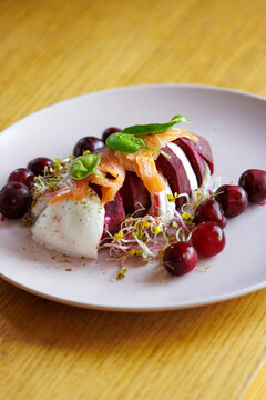Pieces of burrata cheese interspersed with pieces of red beetroot, sprinkled on top with sprouts, basil and sliced ​​salmon on a plate with sprigs, a light summer dish.