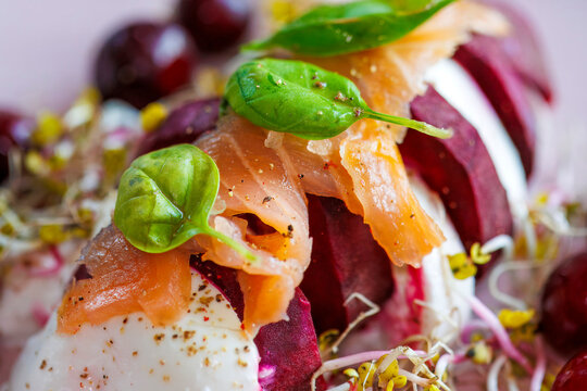 Pieces of burrata cheese interspersed with pieces of red beetroot, sprinkled on top with sprouts, basil and sliced ​​salmon on a plate with sprigs, a light summer dish.