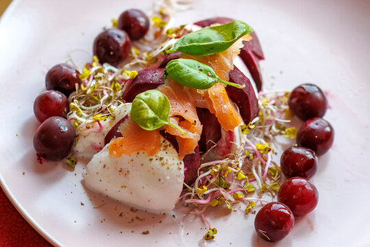 Pieces of burrata cheese interspersed with pieces of red beetroot, sprinkled on top with sprouts, basil and sliced ​​salmon on a plate with sprigs, a light summer dish.