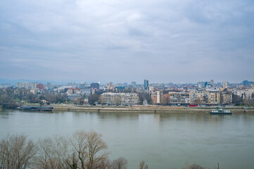 Obraz premium Panoramic view of a city skyline along a river under a cloudy sky in Novi Sad, Serbia, with waterfront buildings and foreground trees.