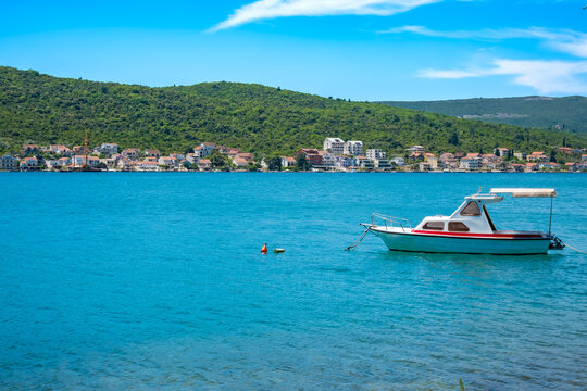 A small boat floats on clear blue water near a coastal town. Lush green hills rise in the background under a bright blue sky.