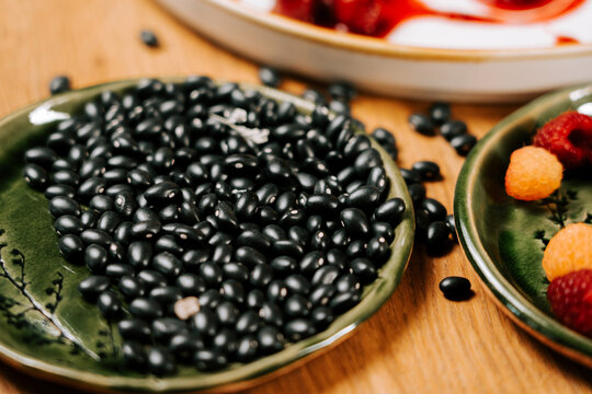 Small black beans arranged on a green decorative plate alongside fresh raspberries on a wooden table