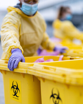 Worker in yellow suit handles hazardous waste bins. Gloves and mask shield against biological risks. Yellow containers mark contamination zones clearly