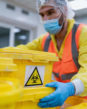Man in protective gear handles hazardous waste container. Yellow bin bears biohazard symbol. He wears mask, gloves, and cap. Focused expression shows responsibility. Ideal for safety, medical