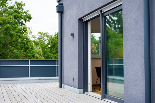 Sliding glass door stands open between composite deck terrace and modern apartment interior with gray stucco facade, metal railing, and green trees surround empty rooftop terrace under overcast sky