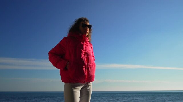 Woman ocean pink jacket lady standing on the coast during a sunny day wearing a bright puffer coat and sunglasses