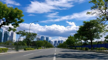 Fototapeta premium Empty road with trees on sides, leading to distant skyscrapers under bright blue sky with fluffy clouds