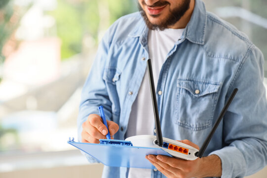 Man in denim shirt holding a wifi router and clipboard, writing notes for internet service installation or repair