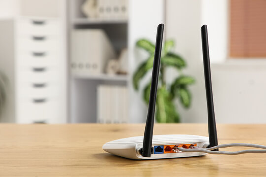 White wireless router with two antennas and connected ethernet cables on a wooden desk in an office setting, the background shows a blurred office environment with a green plant, shelves