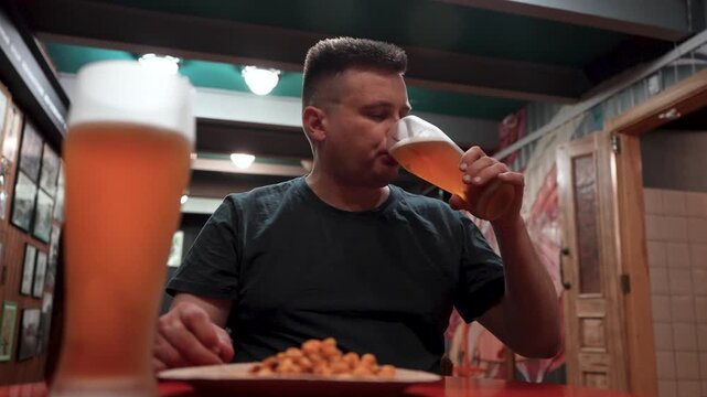 Tired man in a dark T-shirt drinking beer from large glass mug with white foam, sitting at a bar table.