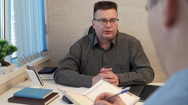Employee writing down tasks in notebook as boss dictates, office instruction and task recording.
